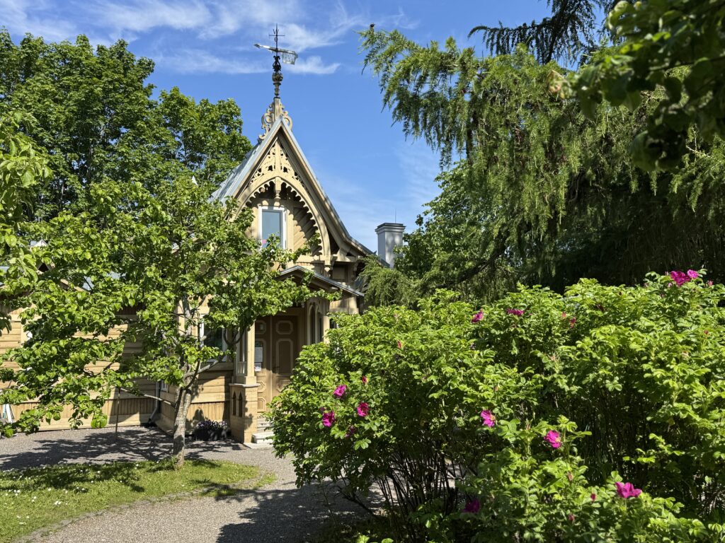 Õuevaade roheliste põõsaste ja puudega päikesepaistelisel päeval, mille vahelt on näha beeži puidust maja esiuks ja dekoratsioonidega katusehari.