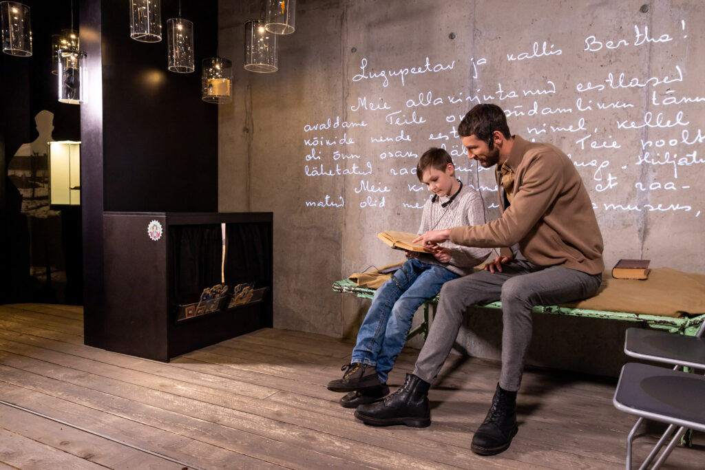 Isa koos pojaga istumas voodi peal ja vaatamas raamatut. Taustaks on Vabamu muuseumi teemaruum "Ebainimlikkus"