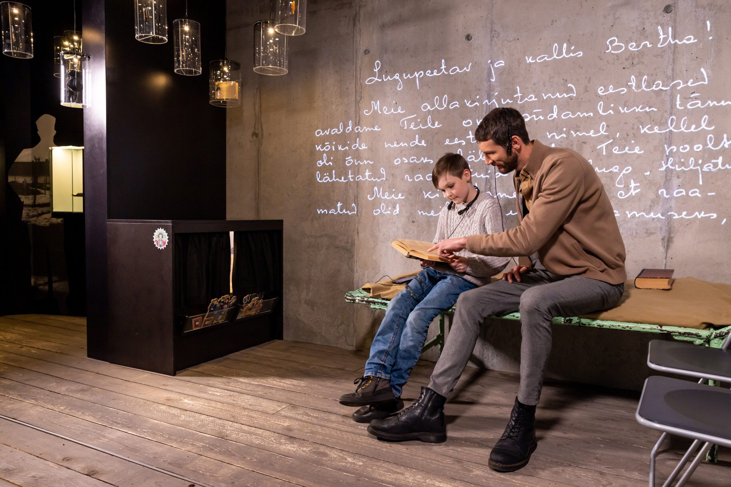 Isa koos pojaga istumas voodi peal ja vaatamas raamatut. Taustaks on Vabamu muuseumi teemaruum "Ebainimlikkus"
